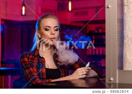 Young pretty woman in a shirt in a cage smoke an electronic cigarette at the vape bar 98062654