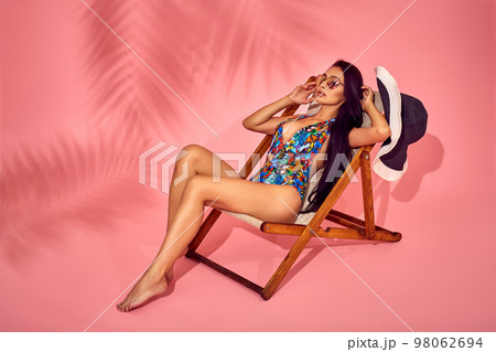 Summer lifestyle fashion portrait of young stunning woman on a lounge chair, pink background, studio shot. Wearing stylish sunglasses, bikini. Summer lifestyle fashion portrait of young stunning woman on a lounge chair, pink background, studio shot. Wearing stylish sunglasses, bikini. 98062694