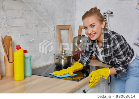 Blonde woman in protective gloves with rag cleaning electric stove at home kitchen. Girl washing black shiny surface of kitchen top Blonde woman in protective gloves with rag cleaning electric stove at home kitchen. Girl washing black shiny surface of kitchen top 98063614