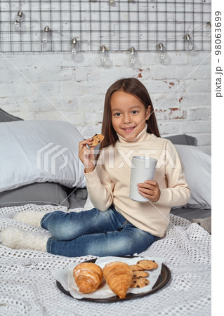 Pretty little girl 6 years old in a white sweater and jeans. Child in the room with a bed, eating croissant and drinking tea or cocoa. Pretty little girl 6 years old in a white sweater and jeans. Child in the room with a bed, eating croissant and drinking tea or cocoa. 98063699