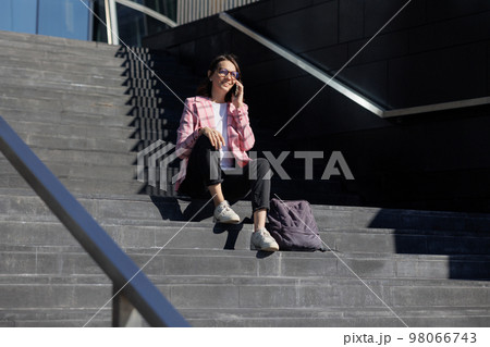business woman in pink jacket is sitting on steps next to office and talking on phone 98066743