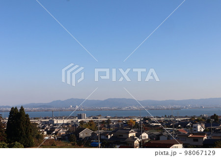 高台からの青空と白む空と市街地の町並みと琵琶湖と対岸の街並みと霞む山並みの風景 98067129