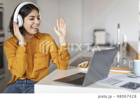Young Arab Female Wearing Headphones Making Video Call On Laptop In Kitchen Young Arab Female Wearing Headphones Making Video Call On Laptop In Kitchen 98067428