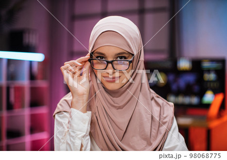 Smiling positive confident woman in formal wear and hijab and eyeglasses sitting at office desk 98068775