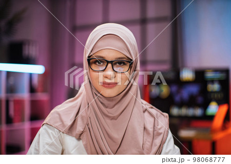 Smiling positive confident woman in formal wear and hijab and eyeglasses sitting at office desk 98068777