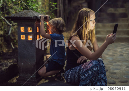 Mom looks into her smartphone, the son looks into the luminous cozy window of the house. The boy lacks Attention, care, love and home comfort 98070911