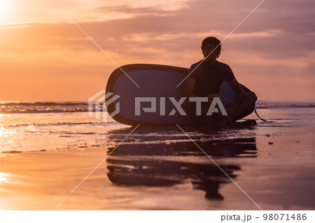 Silhouette of surf man sitting with a surfboard on the seashore beach at sunset time 98071486