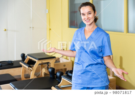 Smiling female physiotherapist inviting to rehab center with pilates equipment Smiling female physiotherapist inviting to rehab center with pilates equipment 98071817
