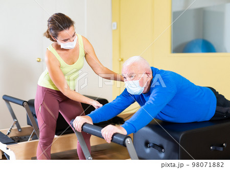 Senior man in protective mask exercising on Pilates reformer with female trainer 98071882
