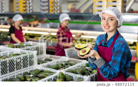Smiling female worker in fruit sorting warehouse holding sliced avocado 98072115