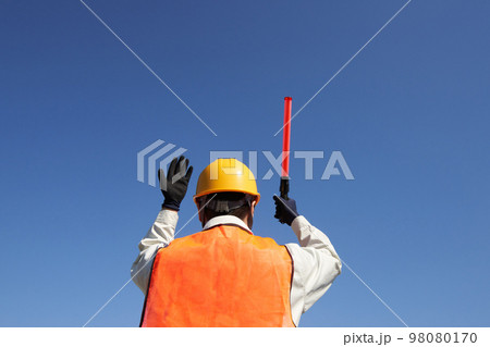 青空のもと交通誘導をする安全ベスト着用の男性の後ろ姿 98080170