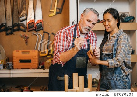 Craftsmen senior man teaching woman apprentice at woodshop, Carpenter training wood for female standing at wood table in workshop, National Carpenters Day Craftsmen senior man teaching woman apprentice at woodshop, Carpenter training wood for female standing at wood table in workshop, National Carpenters Day 98080204