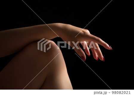 Woman's hand lies relaxed on bare knee on her leg. Crop closeup shot of relaxed female fingers with red nails on black background. Selective and soft focus on human hand. Woman's hand lies relaxed on bare knee on her leg. Crop closeup shot of relaxed female fingers with red nails on black background. Selective and soft focus on human hand. 98082021