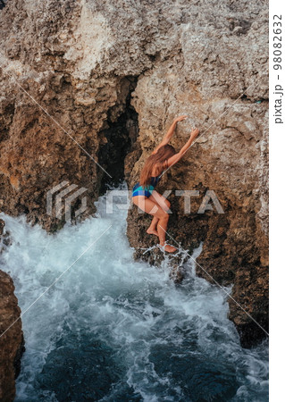 A young beautiful tanned woman with long hair in blue bikini sitting at rocks near blue green clear ocean sea water. Woman relax on sunset at sea, enjoy holidays and weekend vacation in summer time. A young beautiful tanned woman with long hair in blue bikini sitting at rocks near blue green clear ocean sea water. Woman relax on sunset at sea, enjoy holidays and weekend vacation in summer time. 98082632