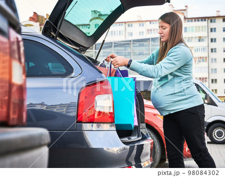 Side view of Caucasian woman with belly puting shopping bags in automobile trunk enjoying maternity and parenthood in awaiting mode. Pregnant female preparing for baby birth. 98084302