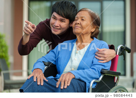 happy senior woman sitting in wheelchair with young man grandson at outside house happy senior woman sitting in wheelchair with young man grandson at outside house 98084443