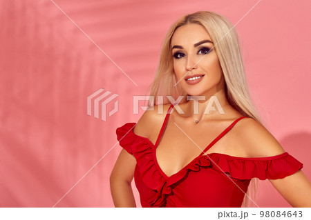 Stunning blonde female model with amazing body standing in an elegant red swimsuit on pink background with palm shadow 98084643