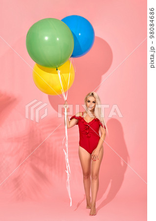 Young sexy slim woman in a red swimsuit with balloons in her hand is posing in a studio. Young sexy slim woman in a red swimsuit with balloons in her hand is posing in a studio. 98084686
