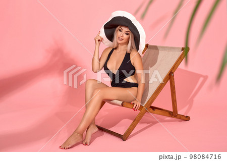 Young woman in black swimsuit relaxing on chaise longue, studio shot. Blurred palm tree leaves in foreground. Young woman in black swimsuit relaxing on chaise longue, studio shot. Blurred palm tree leaves in foreground. 98084716