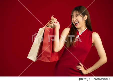 Chinese New Year. Happy asian woman in red chinese dress costume smiling and holding paper shopping bags on red background.. Chinese New Year. Happy asian woman in red chinese dress costume smiling and holding paper shopping bags on red background.. 98089061