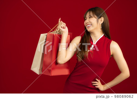 Chinese New Year. Young beautiful asian woman in red chinese dress costume smiling and holding paper shopping bags on red background.. Chinese New Year. Young beautiful asian woman in red chinese dress costume smiling and holding paper shopping bags on red background.. 98089062