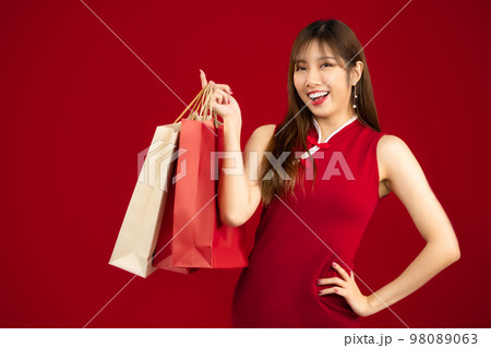 Young beautiful asian woman in red chinese dress costume smiling and holding paper shopping bags on red background.. 98089063
