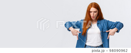 Wait a minute what is that. Frustrated and angry tensed redhead girlfriend asking wtf going on, dont understand, looking perplexed and mad down, pointing bottom copy space, stand white background 98090962