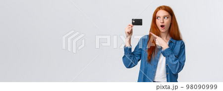 Discounts, shopping and banking concept. Waist-up impressed, thrilled cute redhead girl in denim shirt holding credit card, pointing at it with fascinated impressed eyes, standing white background 98090999