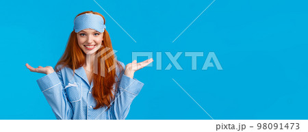 Who knows, dont care. Carefree and unbothered happy smiling redhead female in nightwear, sleep mask, shrugging indecisive and clueless with hands raised sideways, standing blue background 98091473