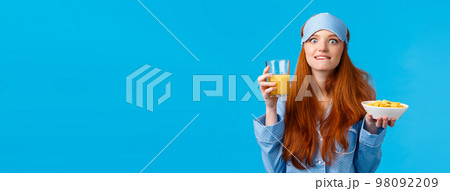 Delicious. Woman eating favorite food in morning. Cute redhead college girl in sleep mask and nightwear, licking lips from temptation and satisfaction, eating tasty cereals and orange juice Delicious. Woman eating favorite food in morning. Cute redhead college girl in sleep mask and nightwear, licking lips from temptation and satisfaction, eating tasty cereals and orange juice 98092209