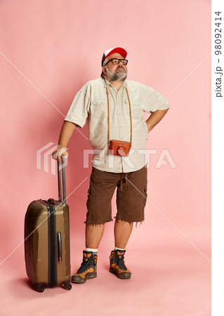 Portrait of mature overweight man in casual clothes with suitcase and vintage camera posing over pink studio background. Tourist Portrait of mature overweight man in casual clothes with suitcase and vintage camera posing over pink studio background. Tourist 98092414