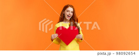 Hey gorgeous you have my heart. Valentines day, boyfriend and girlfriend concept. Cute and sassy redhead woman holding large sign and smiling, wink coquettish, standing orange background 98092654