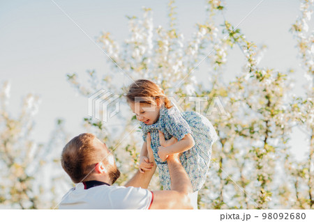 Dad plays and tosses his little daughter on a walk in a blooming spring garden. Dad plays and tosses his little daughter on a walk in a blooming spring garden. 98092680