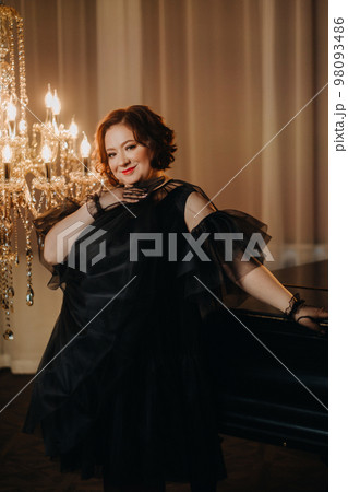 An elegant adult woman in a black cocktail dress poses in the interior. Fashionable portrait of a woman An elegant adult woman in a black cocktail dress poses in the interior. Fashionable portrait of a woman 98093486