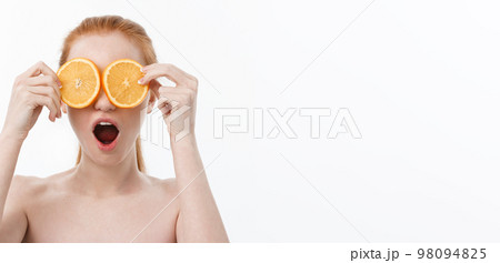 Portrait of a cheerful young girl holding two slices of an orange at her face over white wall background Portrait of a cheerful young girl holding two slices of an orange at her face over white wall background 98094825