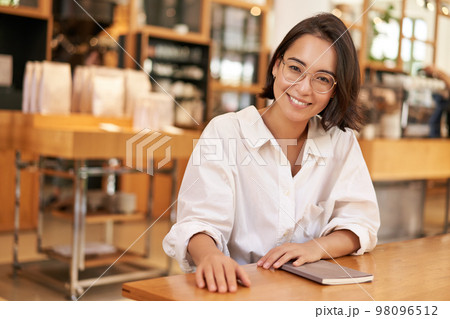 Portrait of brunette woman in glasses, smiling with confident look, sitting in cafe with notebook, working from restaurant 98096512