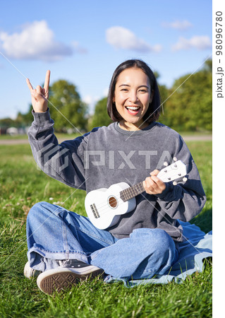 Happy asian girl playing ukulele in park, showing rock n roll, heavy metal horns sign and smiling, having fun outdoors Happy asian girl playing ukulele in park, showing rock n roll, heavy metal horns sign and smiling, having fun outdoors 98096780
