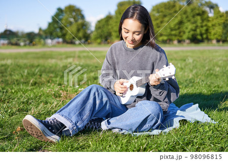 Carefree asian girl singing and playing ukulele in park, sitting on grass, musician relaxing on her free time outdoors Carefree asian girl singing and playing ukulele in park, sitting on grass, musician relaxing on her free time outdoors 98096815