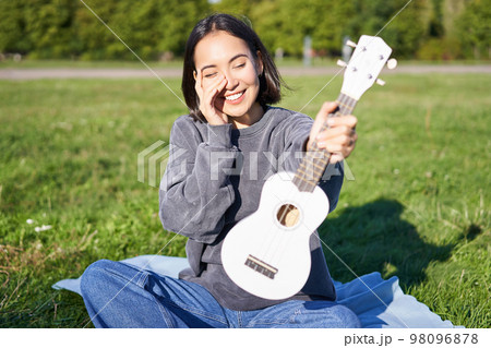 Beautiful asian girl with happy smile, shows her ukulele, sits outside in park on grass, relaxes with music Beautiful asian girl with happy smile, shows her ukulele, sits outside in park on grass, relaxes with music 98096878