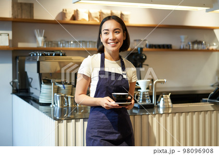 Portrait of asian smiling female barista, cafe waitress holding cup of coffee, serving clients, taking order to the tablet, standing near cafe counter 98096908