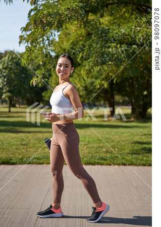 Sport and wellbeing. Young sportswoman in park, listen music and smiling, workout outdoors, jogging on streets of city 98097078