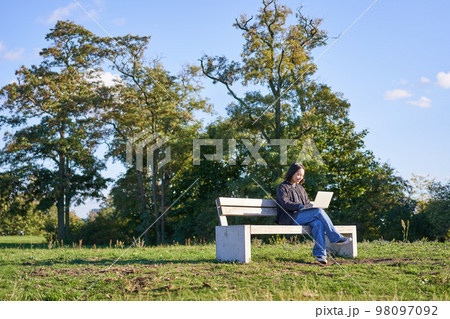 Portrait of young student, girl using laptop, sitting in park on bench, typing on computer 98097092