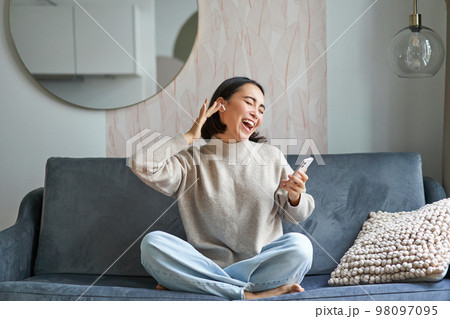 Portrait of carefree asian woman singing and listening music from smartphone app, using wireless headphones, smiling pleased, sitting on sofa at home Portrait of carefree asian woman singing and listening music from smartphone app, using wireless headphones, smiling pleased, sitting on sofa at home 98097095