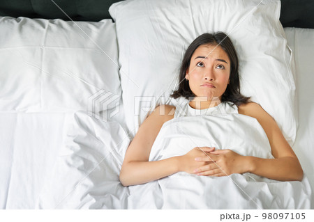 Portrait of asian girl lying in her bed and shrugs, looks puzzled, overthinking before going to sleep, looking complicated at camera 98097105