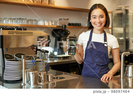 Smiling asian girl barista, cafe owner in apron, showing card machine, payment reader, taking contactless orders in her coffee shop 98097464
