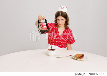 Stylish young female waitress in retro american fashion style of 70s, 80s sitting at table and drinking coffee over grey background. Funny meme emotions, ad, sales Stylish young female waitress in retro american fashion style of 70s, 80s sitting at table and drinking coffee over grey background. Funny meme emotions, ad, sales 98097633