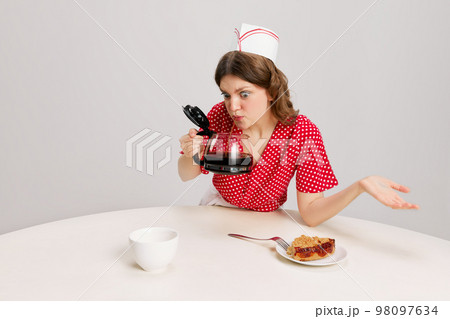 One young woman, strange waitress in 70s, 80s retro fashion style uniform sitting at table with coffee over grey background. Funny meme emotions, ad, sales 98097634