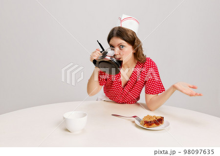 Stylish young female waitress in retro american fashion style of 70s, 80s sitting at table and drinking coffee over grey background. Funny meme emotions, ad, sales Stylish young female waitress in retro american fashion style of 70s, 80s sitting at table and drinking coffee over grey background. Funny meme emotions, ad, sales 98097635