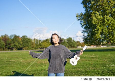Upbeat young woman dancing with her musical instrument. Girl raises her ukulele up and pose in park on green field 98097641