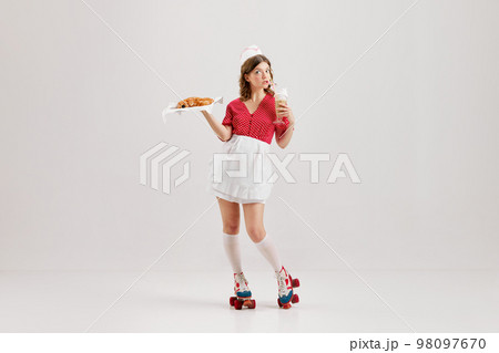 Emotional young woman, retro waitress in american fashion style of 70s, 80s uniform posing over light studio background. Comic photography style 98097670